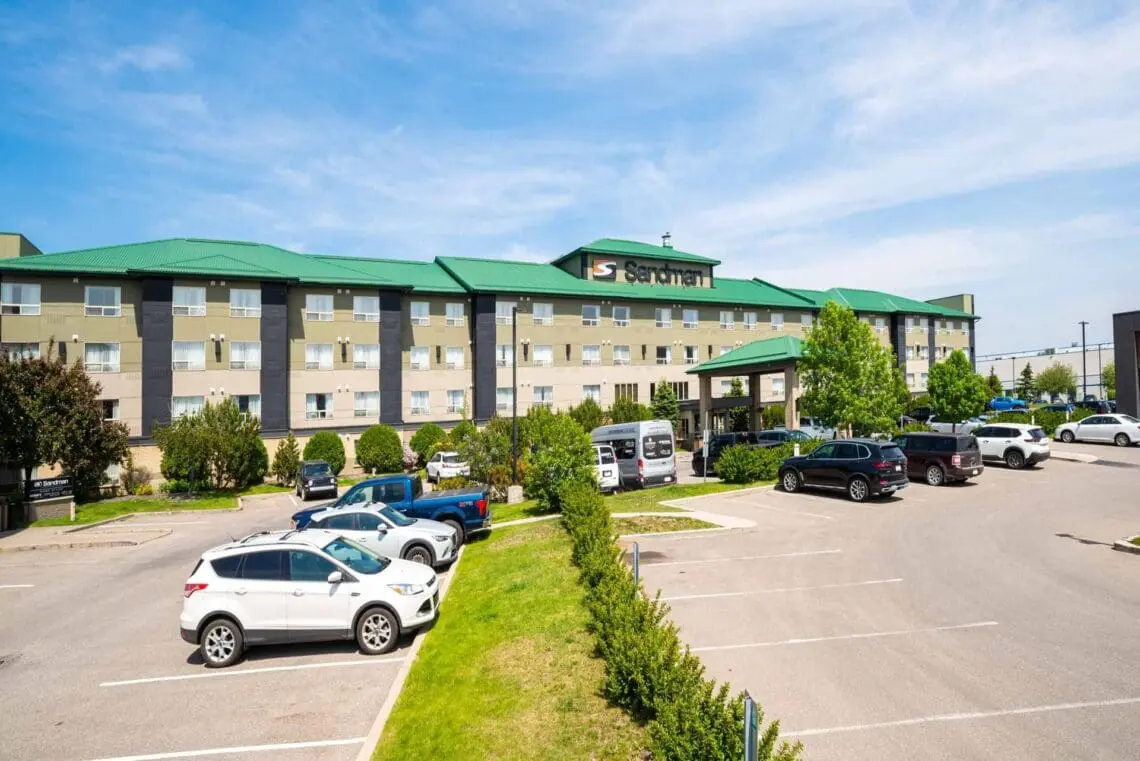 A hotel building with a green roof and sign reading "Sandman" rises behind a parking lot filled with cars under a partly cloudy sky, subtly elevating commercial landscapes with its inviting presence.