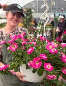 Annual Team 2 A person wearing a cap and apron holds a white hanging pot filled with bright pink and white flowers inside an annual greenhouse.