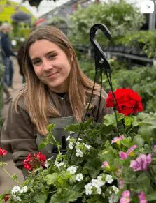 Annual Team A young woman with straight brown hair smiles beside a hanging basket of colorful flowers in an annual greenhouse or garden center.