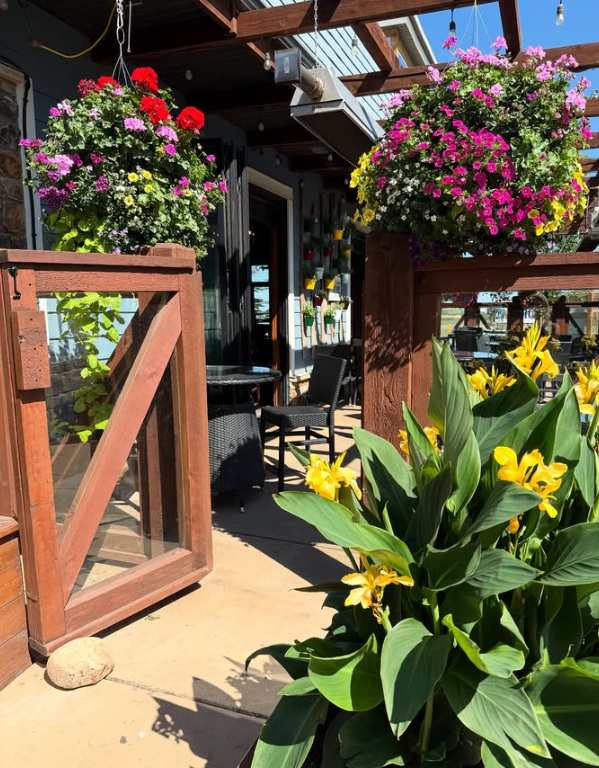 Kildares Outdoor patio with wooden gate, hanging baskets of colorful flowers from an annual greenhouse, yellow flowers in the foreground, and patio seating in the background under a pergola.