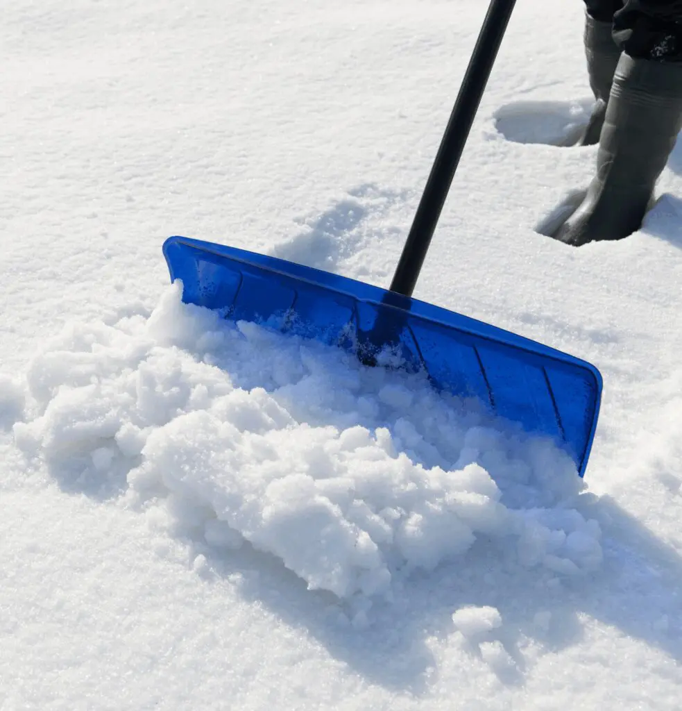 Person using a blue snow shovel to clear fresh snow, with only their legs and boots visible in the background.