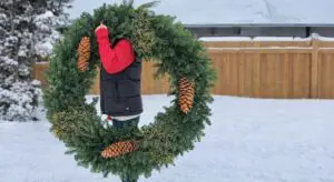 A person in a red jacket holds a large evergreen wreath with pine cones, standing outside in a snowy yard near a wooden fence.