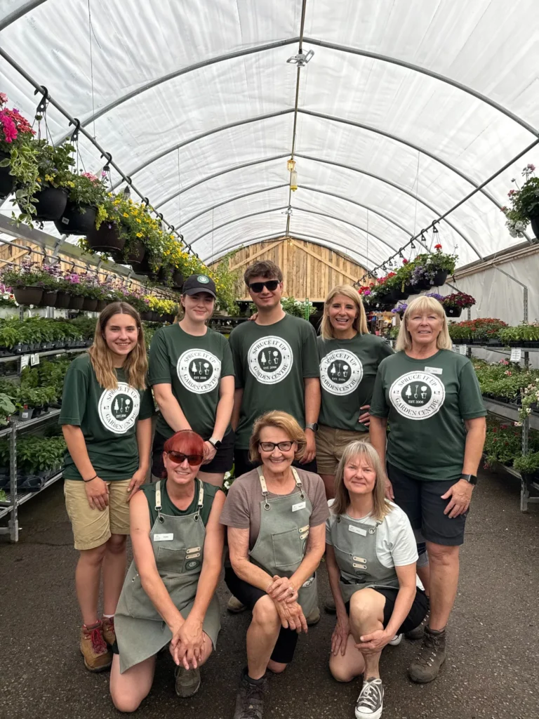 Eight people pose inside the Annual Greenhouse, wearing matching green shirts with a logo and name tags. Flowering plants and lush greenery fill the surrounding shelves.