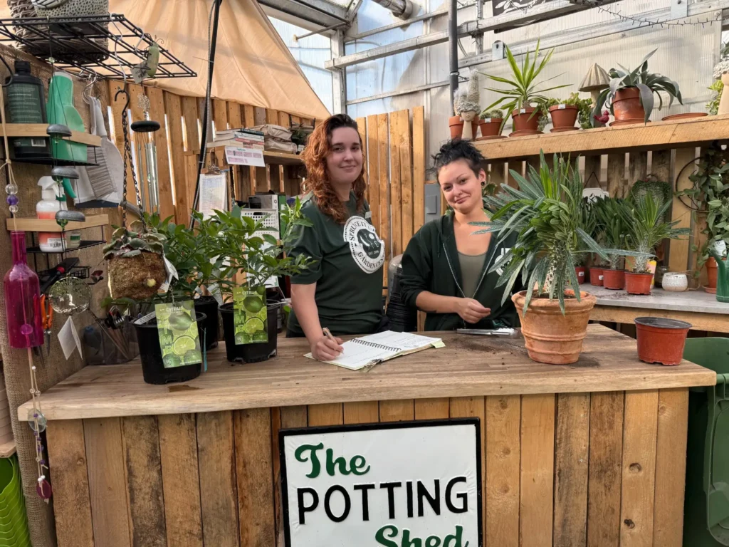 Potting_Station2_compressed Two women stand behind a wooden counter in a greenhouse, surrounded by potted plants and gardening tools, with a sign reading