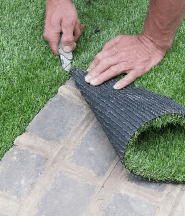 A person uses a utility knife to trim a section of artificial grass next to a paved surface.