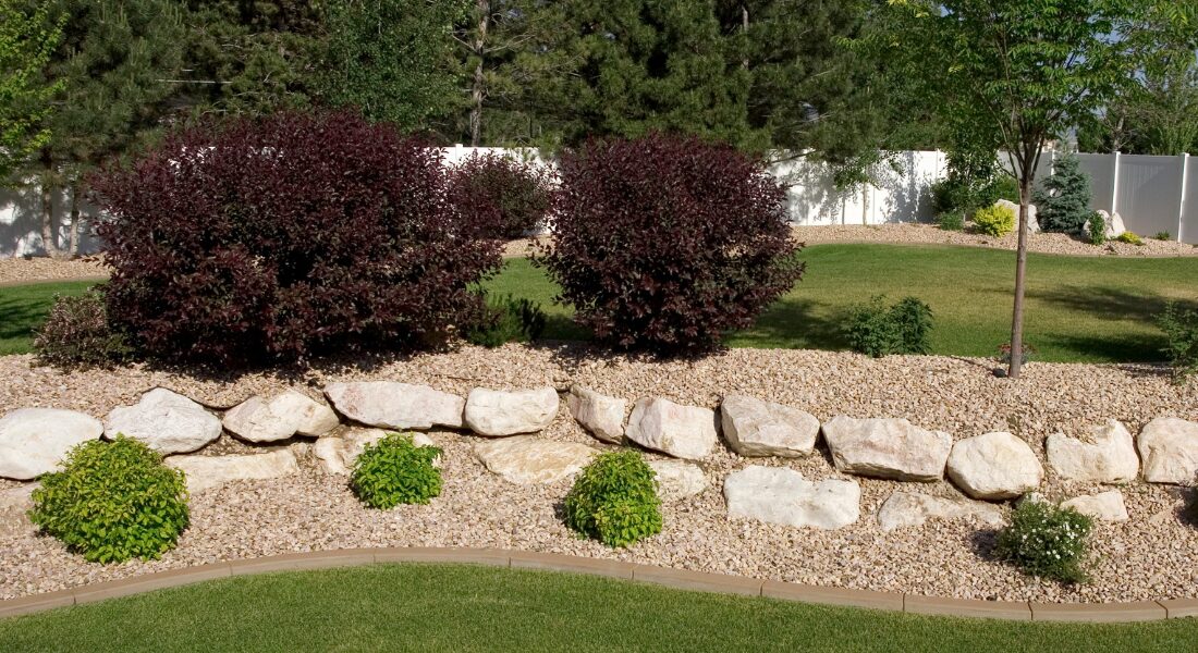 A landscaped backyard features a stone border, gravel, shrubs, green grass, and trees with a white fence in the background.